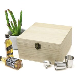 May include: A wooden box with a brass latch, two small bottles of liquor, a funnel, and two shot glasses. The box is light brown and has a natural wood grain. The bottles are clear glass with black labels. The funnel is silver metal. The shot glasses are silver metal.