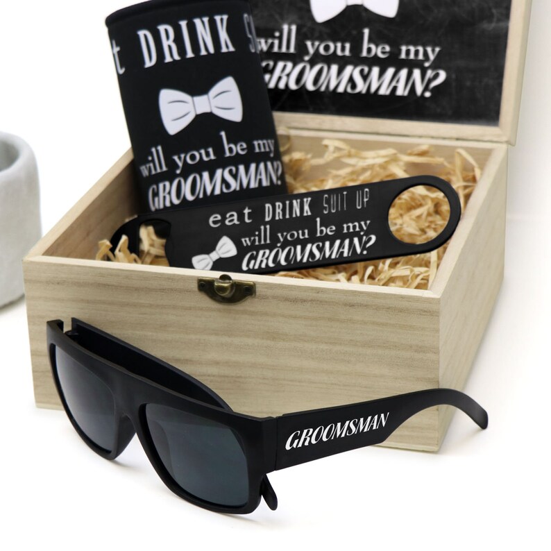 May include: A wooden box filled with a black can cooler, a black bottle opener, and black sunglasses. The can cooler and bottle opener have white text that says "Will you be my groomsman?" and "Eat, Drink, Suit Up". The sunglasses have white text that says "Groomsman".