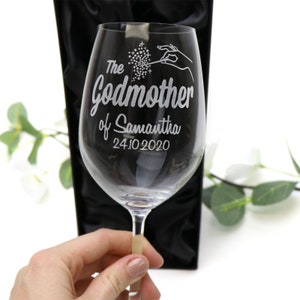 May include: A clear glass wine glass with an etched design of a hand holding a wand with sparkles. The text on the glass reads "The Godmother of Samantha 24.10.2020".