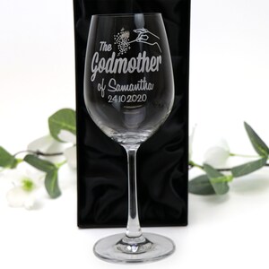May include: A clear glass wine glass with an engraved design that says "The Godmother of Samantha 24.10.2020". The glass is in a black box with a satin lining.