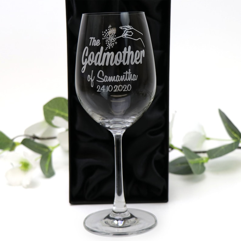May include: A clear glass wine glass with an engraved design that says "The Godmother of Samantha 24.10.2020". The glass is in a black box with a satin lining.