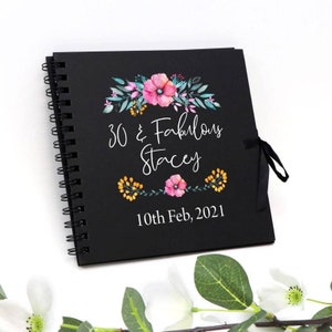 May include: A black spiral-bound journal with the text "30 & Fabulous Stacey 10th Feb, 2021" in white script. The cover features floral designs in pink, yellow, and teal. A black ribbon is tied on the right side.