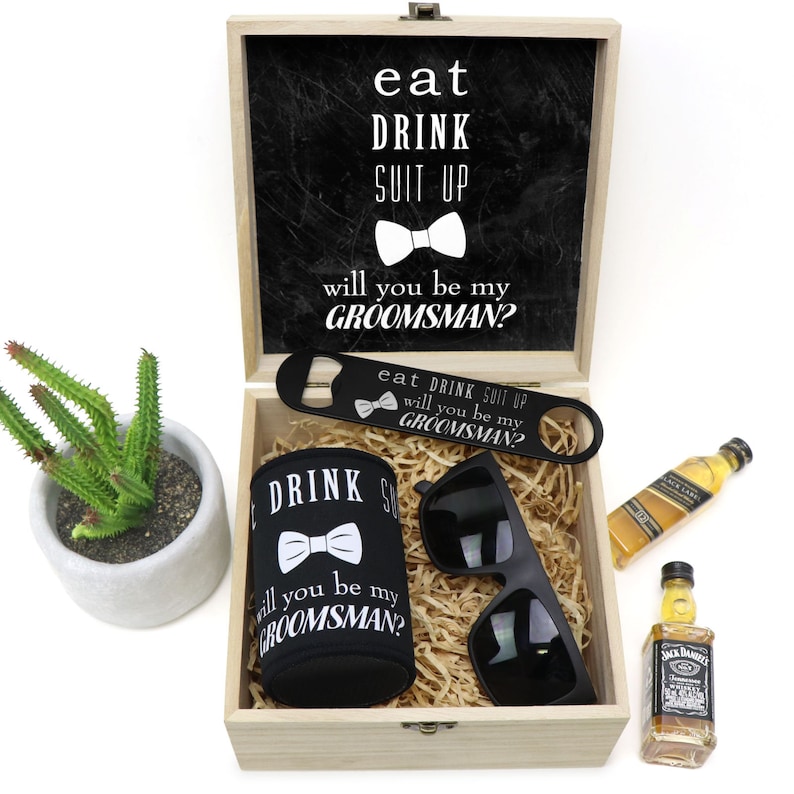 May include: A wooden box with a black chalkboard lid that says "eat drink suit up will you be my groomsman?" The box contains a black can cooler with the same text, a bottle opener, sunglasses, and a miniature bottle of whiskey.