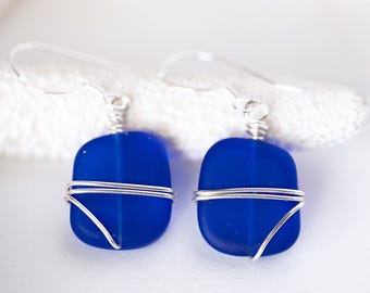 May include: A pair of square, cobalt blue earrings with silver wire detailing. The earrings are suspended from clear, curved ear wires. The earrings are displayed on a white starfish against a white background.