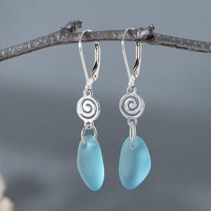 May include: A pair of silver leverback earrings with a spiral design and teardrop-shaped, light blue sea glass pendants. The earrings are displayed on a gray branch against a gray background.