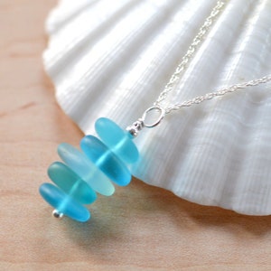 May include: A silver chain necklace with a pendant made of stacked, graduated, light blue sea glass. The pendant is shaped like a teardrop.