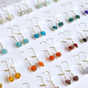 May include: A display of handmade earrings with various colored glass beads. The earrings are silver and have a simple design. The beads are round and have a frosted finish.