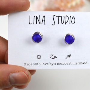 May include: A pair of small, cobalt blue glass earrings with silver wire settings. The earrings are displayed on a white card with the text "LINA STUDIO" and "Made with love by a seacoast mermaid".