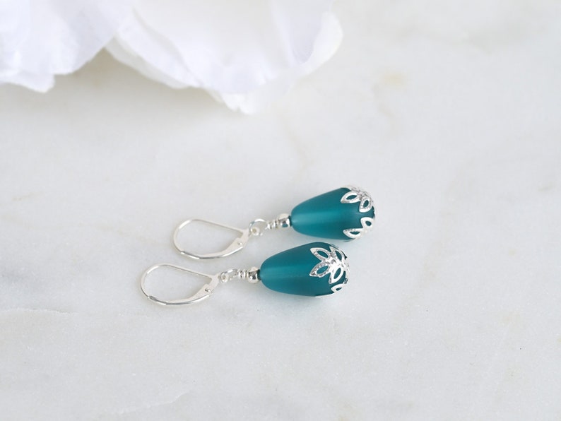 May include: Teal teardrop earrings with silver-colored floral accents. The earrings have a lever-back closure. The background is a white marble surface with a white flower in the upper left corner.