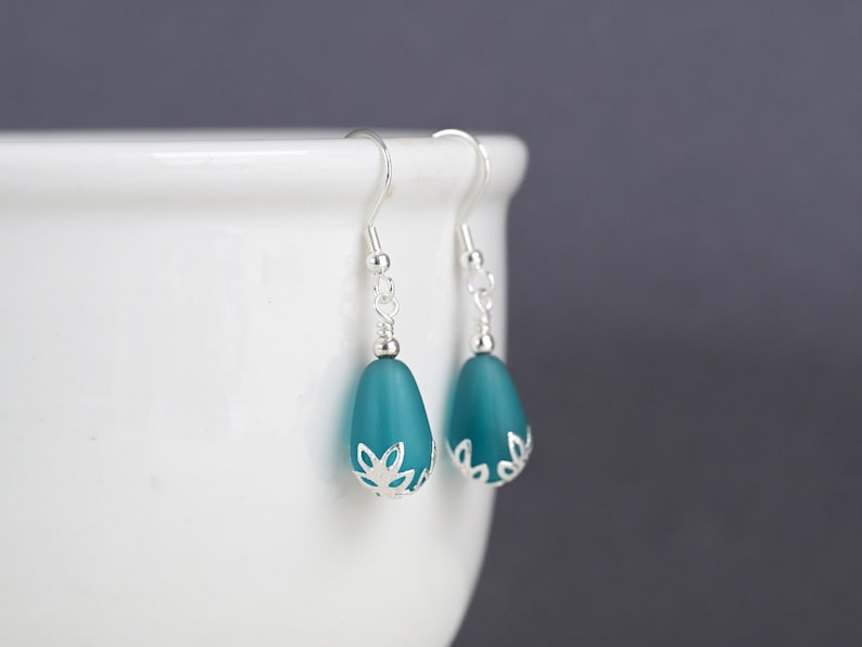 May include: A pair of teal teardrop earrings with silver-colored accents. The earrings have a frosted glass appearance and are suspended from silver-colored hooks. The bottom of each earring features a decorative silver-colored floral design.
