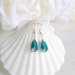 May include: Teal teardrop earrings with silver-colored accents. The earrings have a frosted glass appearance and are displayed on a white seashell. The earrings have a floral design at the base.