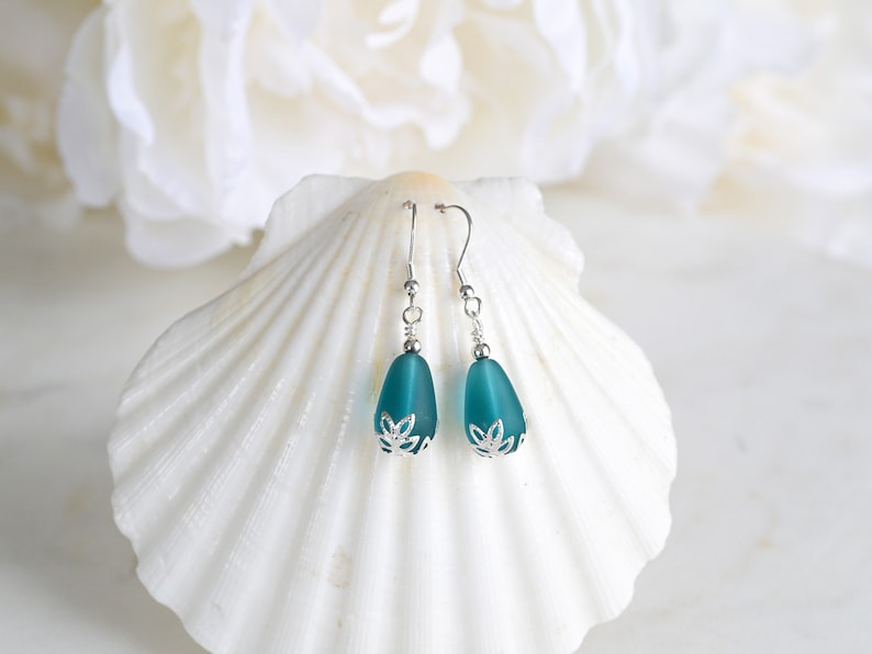 May include: Teal teardrop earrings with silver-colored accents. The earrings have a frosted glass appearance and are displayed on a white seashell. The earrings have a floral design at the base.