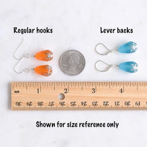 May include: Earrings with regular hooks and lever backs are shown for size reference. The orange earrings have a silver-colored floral design, while the blue earrings also have a silver-colored floral design. A ruler shows the earrings' size in inches.