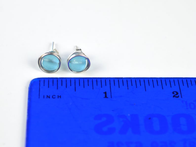 May include: A pair of silver stud earrings with light blue round glass stones. The earrings are shown next to a ruler, measuring approximately 1/2 inch in diameter.