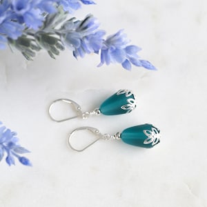 May include: Teal-colored drop earrings with silver-colored floral accents. The earrings have a lever-back closure. The background includes blue flowers and a white marble surface. Jewelry.