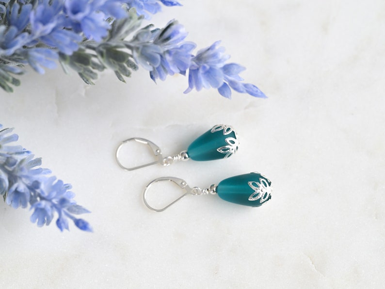 May include: Teal-colored drop earrings with silver-colored floral accents. The earrings have a lever-back closure. The background includes blue flowers and a white marble surface. Jewelry.
