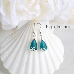 May include: A pair of teal teardrop earrings with silver-colored hooks and decorative caps. The earrings are displayed on a white seashell against a soft, blurred background. The text "Regular hooks" is visible.