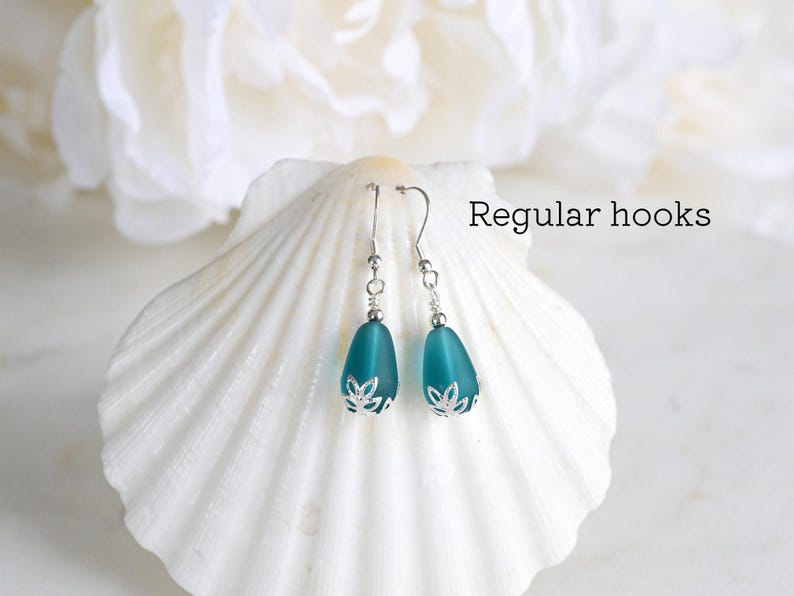 May include: A pair of teal teardrop earrings with silver-colored hooks and decorative caps. The earrings are displayed on a white seashell against a soft, blurred background. The text "Regular hooks" is visible.
