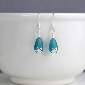 May include: A pair of teal teardrop earrings with silver-colored accents. The earrings feature a frosted glass bead with a decorative silver cap and a simple hook closure. The earrings are displayed against a white background.