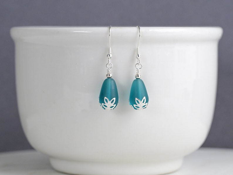 May include: A pair of teal teardrop earrings with silver-colored accents. The earrings feature a frosted glass bead with a decorative silver cap and a simple hook closure. The earrings are displayed against a white background.