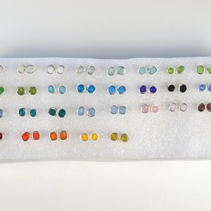 May include: A display of 24 pairs of silver stud earrings with various colored glass stones. The stones are arranged in rows of two, with each row featuring a different color. The colors include white, light blue, green, blue, black, pink, brown, and orange.