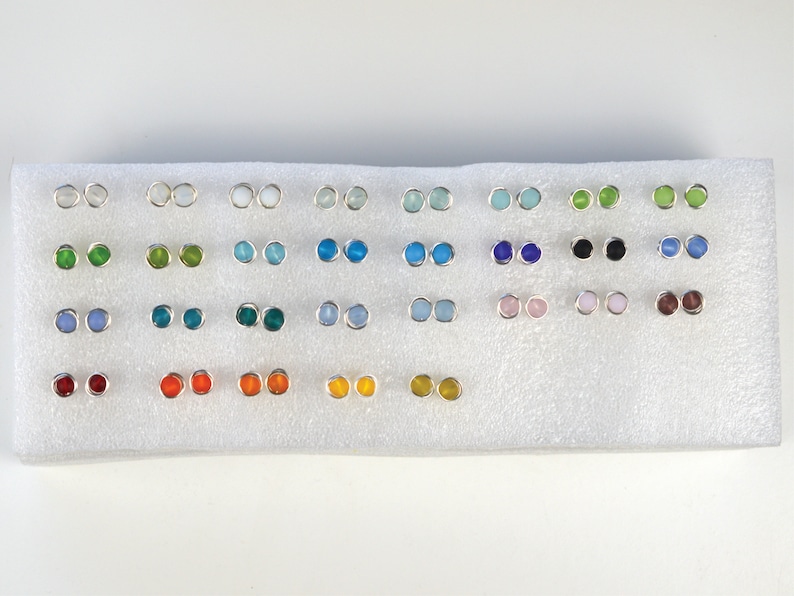 May include: A display of 24 pairs of silver stud earrings with various colored glass stones. The stones are arranged in rows of two, with each row featuring a different color. The colors include white, light blue, green, blue, black, pink, brown, and orange.