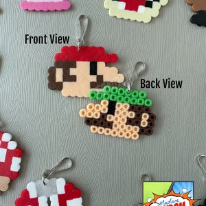 May include: Pixelated Mario keychain, front and back views. The front shows Mario's face with a red hat, brown hair, and a tan face. The back view shows the back of Mario's head with green hair. Other pixelated character keychains are visible.