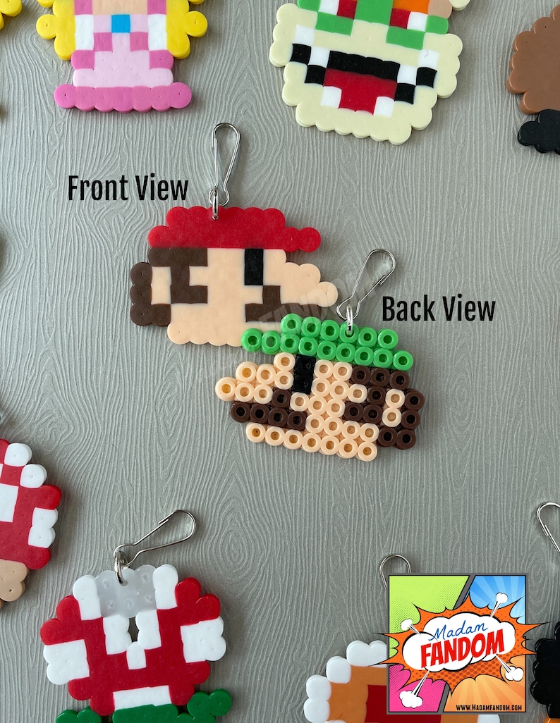 May include: Pixelated Mario keychain, front and back views. The front shows Mario's face with a red hat, brown hair, and a tan face. The back view shows the back of Mario's head with green hair. Other pixelated character keychains are visible.