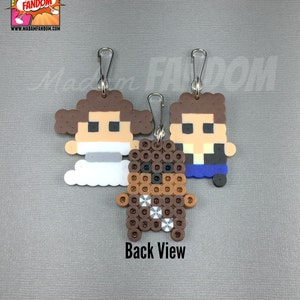 May include: Three pixel art character keychains: Princess Leia, Chewbacca, and Han Solo. The Leia keychain has a white dress and brown hair. Chewbacca is brown with a silver bandolier. Han Solo has a blue jacket. Each keychain has a silver clasp.