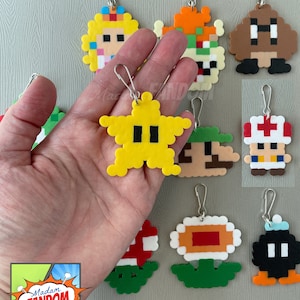 May include: A collection of pixel art keychains featuring characters from the Super Mario video game series. The keychains are made from colorful beads and include characters like Princess Peach, Bowser, and a yellow star. Each keychain has a silver clasp.