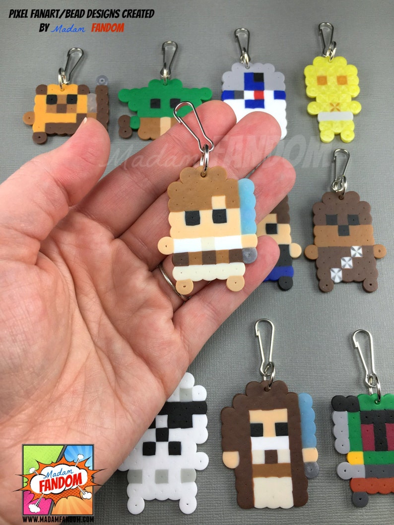 May include: A collection of pixel art keychains featuring various characters. The keychains are made of colorful beads and have silver-colored clasps. The image includes keychains of a character with a lightsaber, a green character, and a yellow character.