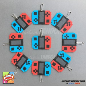May include: A collection of pixel art keychains in the shape of a game console controller. The controllers are red, blue, and gray, with a silver clasp attached. The keychains are arranged in a circle on a gray surface.