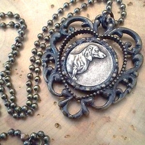 May include: A silver dachshund charm pendant on a brass chain necklace. The pendant is set in a decorative filigree frame.