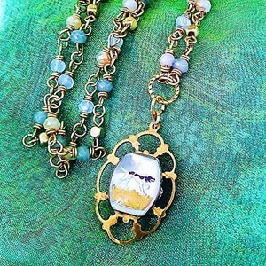 May include: A gold necklace with a pendant featuring a detailed oval frame with a glass cameo of a dog running through a field. The necklace is made with a double strand of gold chain and light blue, white, and pink beads.
