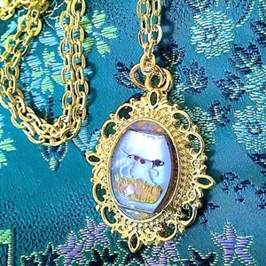 May include: Gold-toned pendant necklace featuring an oval-shaped pendant with a painted dog scene. The pendant is framed by an ornate, filigree-style border. The chain is a classic link style. The background is a teal fabric with a floral pattern.