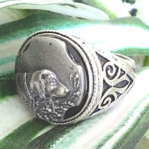 May include: Silver-toned ring featuring a detailed profile of a dog within a shield-shaped design. The ring has an ornate, scrollwork pattern on the sides. The ring is set against a green and white striped background.