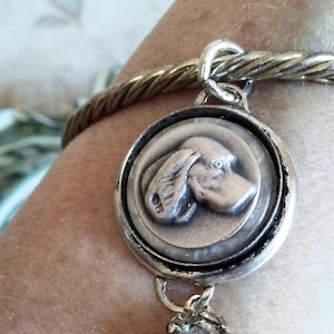May include: Silver-toned bracelet featuring a round pendant with a dog's profile in relief. The pendant has a decorative border and a small, sparkling charm. The bracelet has a twisted metal design.