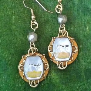 May include: A pair of gold earrings with a white cameo featuring a dog running through a field. The cameo is surrounded by a gold filigree design and the earrings are accented with white beads.