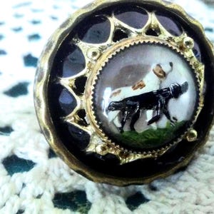 May include: A round decorative button featuring a central circular inlay with a painted illustration of a black dog and a white dog standing on green grass. The inlay is surrounded by a gold-toned metal frame with a scalloped edge and black enamel.