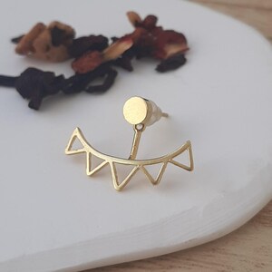 Puede incluir: Pendiente de oreja geométrico dorado con un diseño triangular. El pendiente tiene un pequeño poste redondo que atraviesa la oreja.