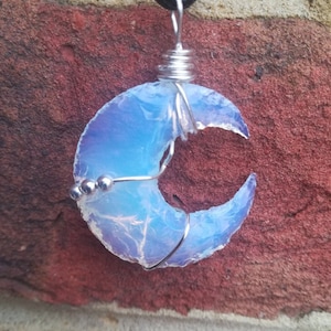 May include: A crescent moon shaped pendant made from iridescent blue opal, wrapped in silver wire with silver beads. The pendant is hanging from a black cord.
