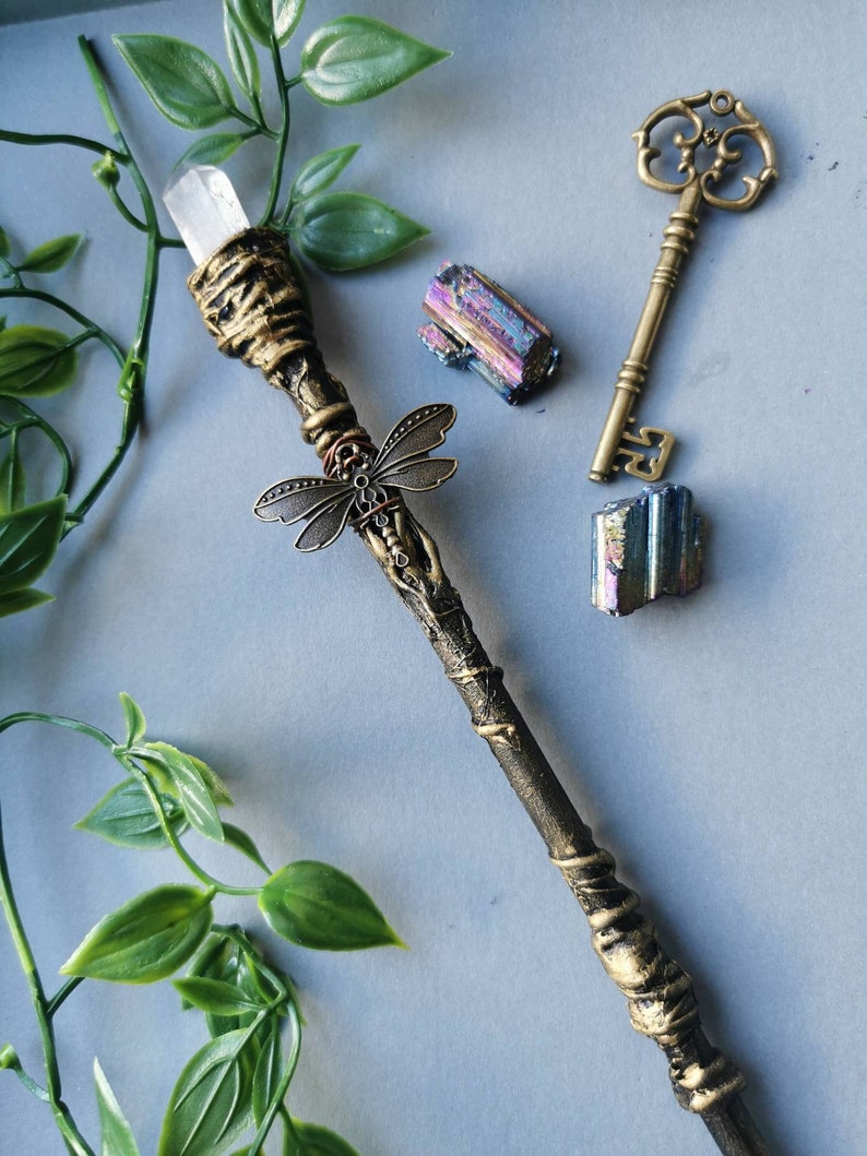May include: A gold and brown wand with a clear crystal at the top and a dragonfly charm. The wand is surrounded by green leaves and two iridescent crystals.
