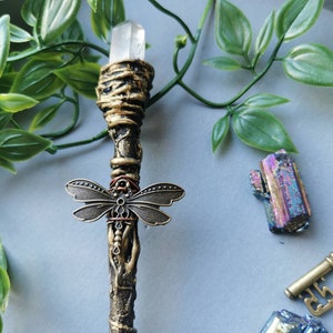 May include: A gold and brown wand with a clear crystal at the top and a dragonfly charm in the middle. The wand is on a gray surface with green leaves and iridescent crystals.