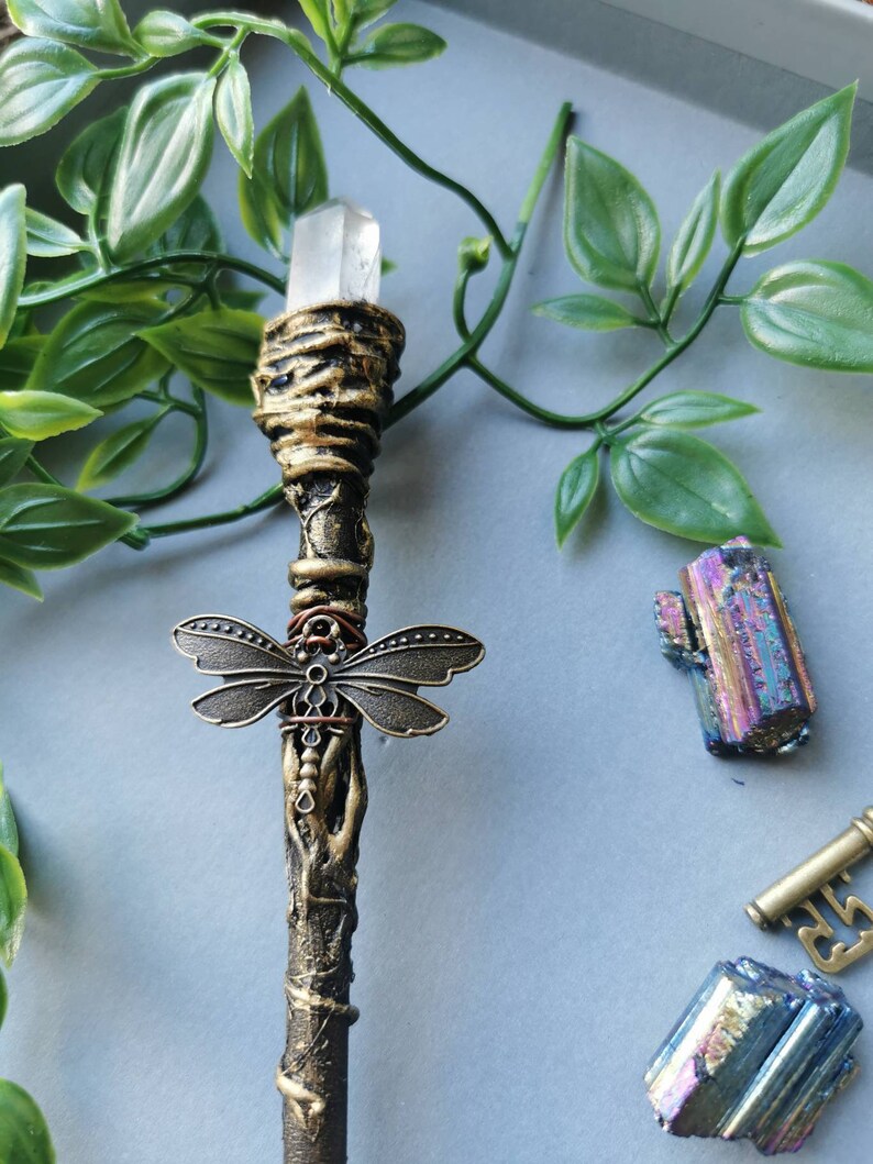 May include: A gold and brown wand with a clear crystal at the top and a dragonfly charm in the middle. The wand is on a gray surface with green leaves and iridescent crystals.