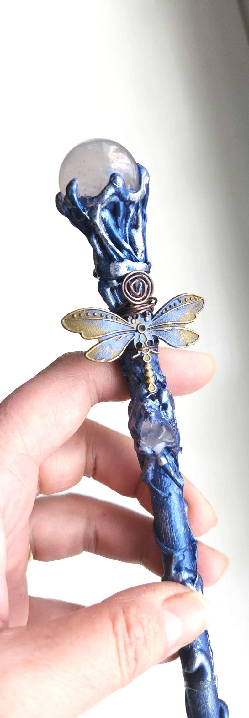 May include: A handcrafted blue wand with a clear crystal orb at the top. The wand features a dragonfly charm with gold and blue wings, and a copper wire spiral. A unique fantasy item.