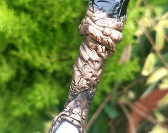 May include: A handcrafted wand with a black obsidian crystal point. The wand features a bronze-colored, intricately carved handle with a white, oval-shaped stone near the base. The background is a blurred green foliage.