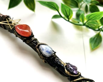 May include: A wooden wand with a gold wire wrapped around it. The wand is decorated with three gemstones: a red carnelian, a blue moonstone, and a purple amethyst. The wand is a magical tool used in witchcraft and pagan rituals.