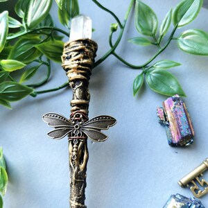 May include: A gold and brown wand with a clear crystal at the top. The wand is decorated with a metal dragonfly and a copper wire wrap. The wand is lying on a light blue surface.