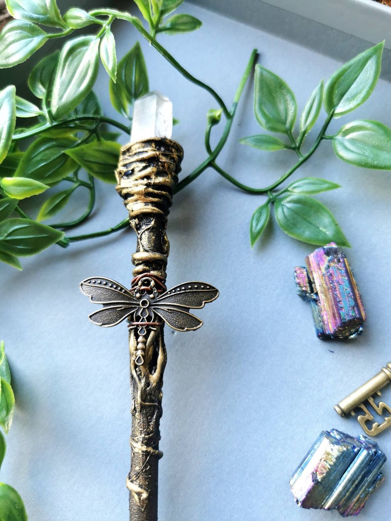 May include: A gold and brown wand with a clear crystal at the top. The wand is decorated with a metal dragonfly and a copper wire wrap. The wand is lying on a light blue surface.