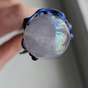 May include: A close-up view of a clear, round, iridescent crystal ball held by a dark blue, textured, claw-like setting. The crystal ball has a subtle, pearly sheen and displays a rainbow of colors. The setting appears to be made of a resin or similar material.